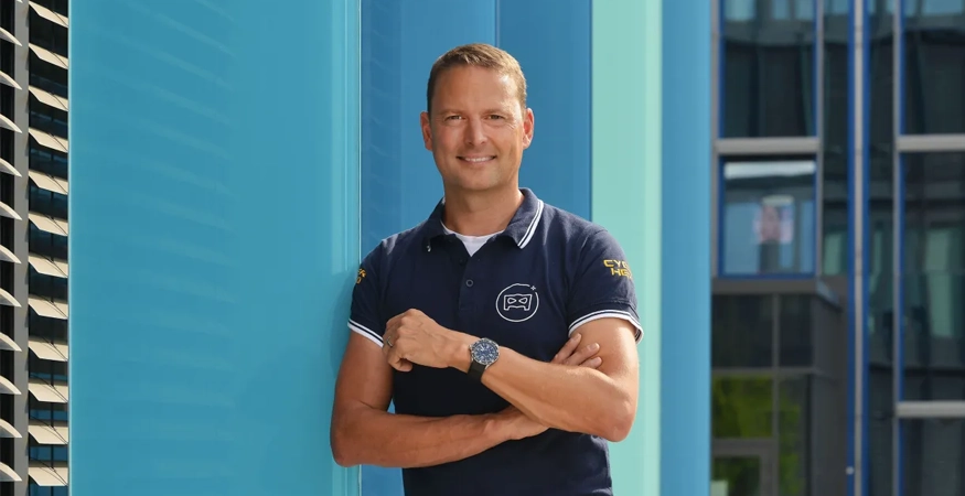 Ein Person ist gegen eine blaue Mauer gelehnt und lächtelt in die Kamera. 