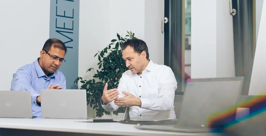 Edward Aston sitzt am Schreibtisch mit einem Kollegen und diskutiert. 